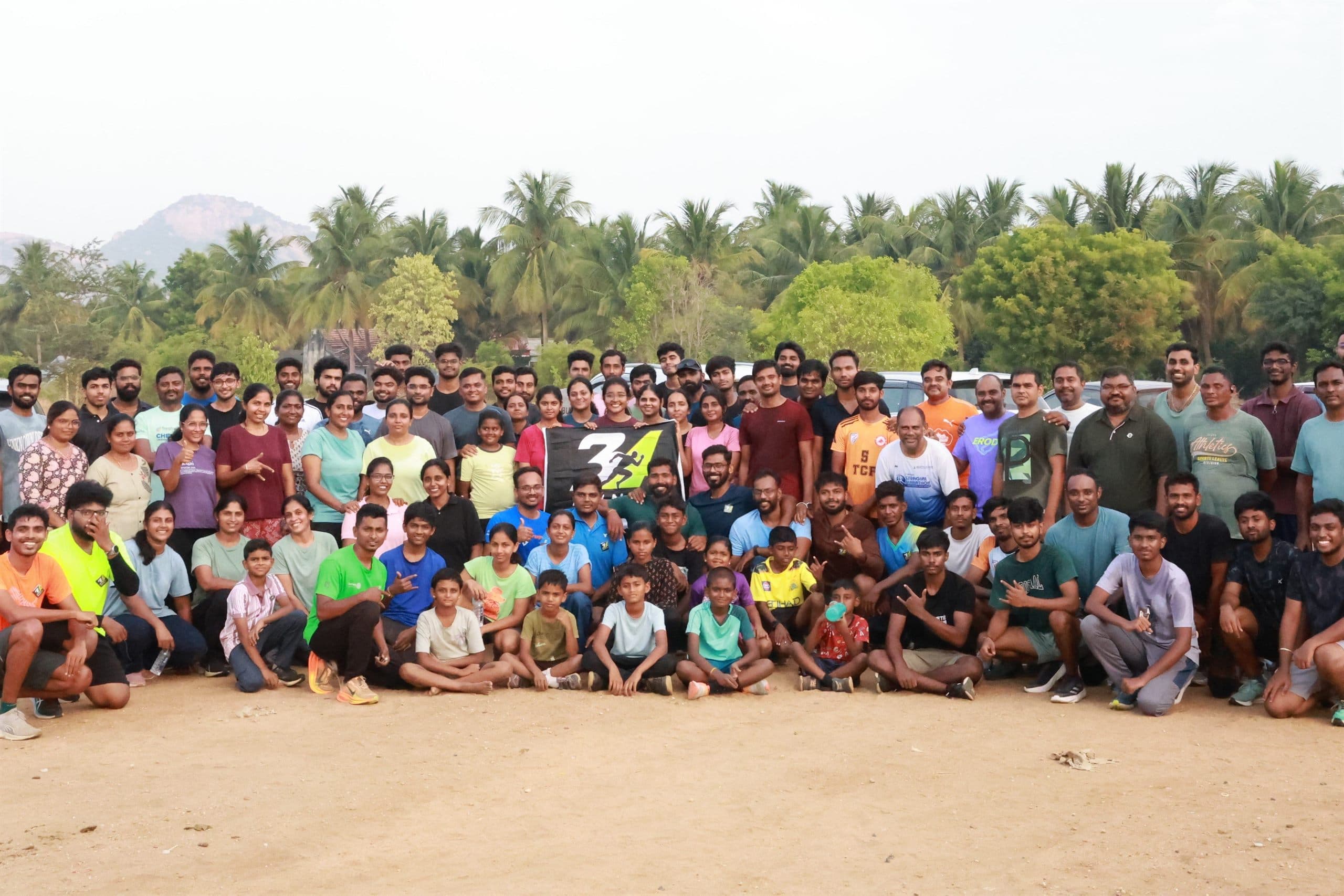 More than a hundred RUN 34 members gathered on open ground for a group photo, standing, kneeling and seated in rows behind the club flag, with palm trees and a hill behind them.