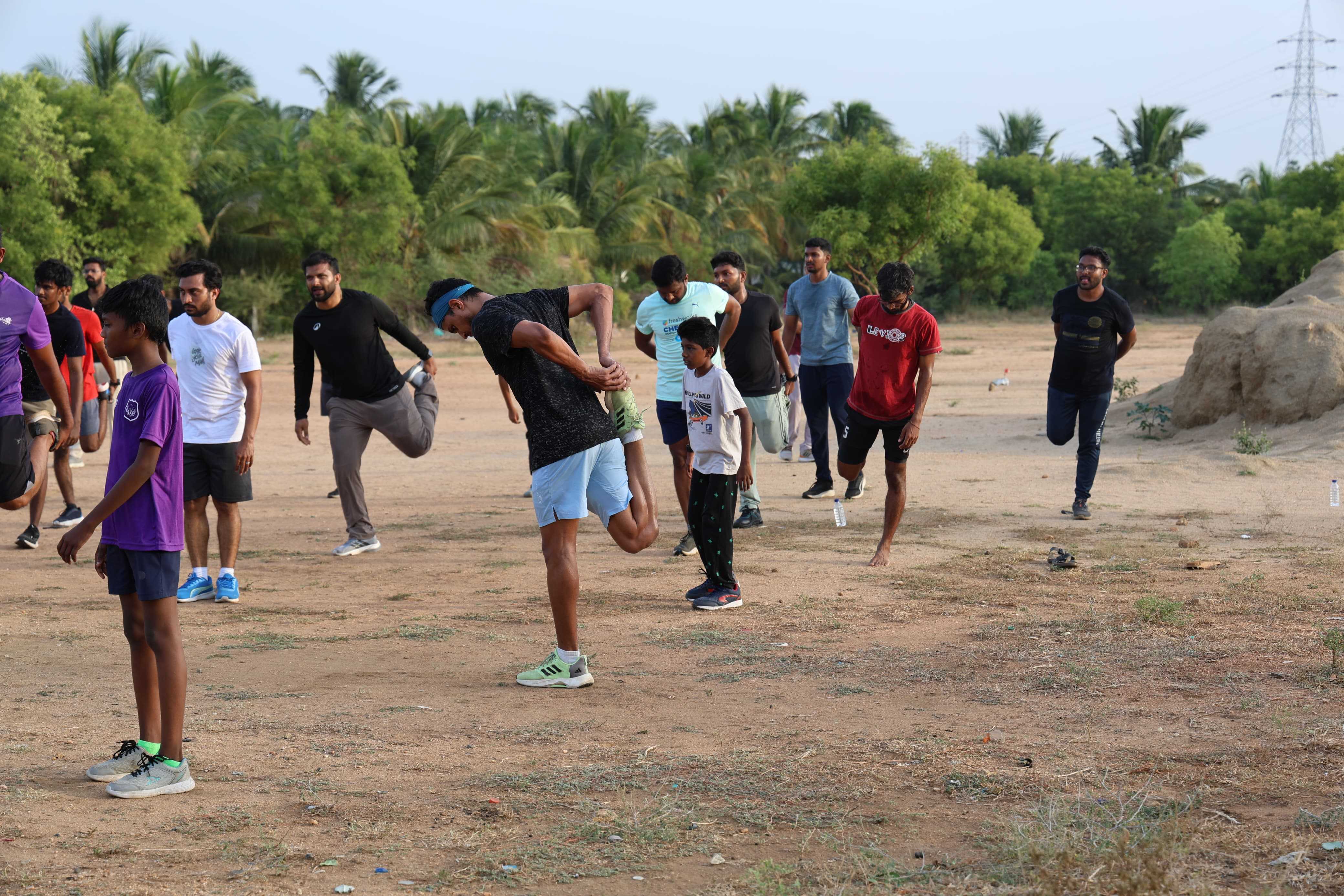 Club members spread across open ground holding quad stretches during the pre-run warm-up, a young runner watching from the foreground.
