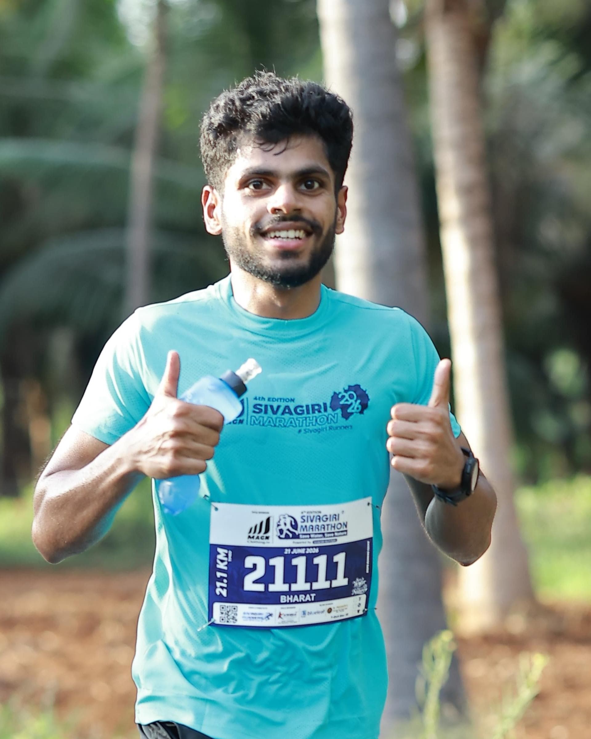 Bharat running a half marathon on a tree-lined road, both thumbs up and smiling at the camera.