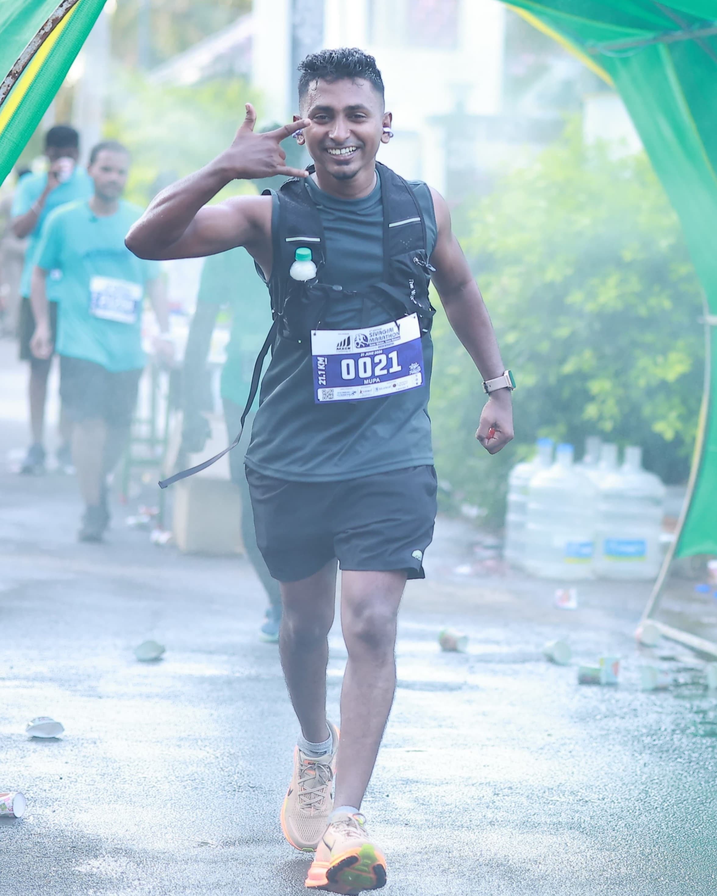 Dinesh mid-run in a black vest and hydration pack, grinning at the camera with one hand raised beside his face, another runner behind him.