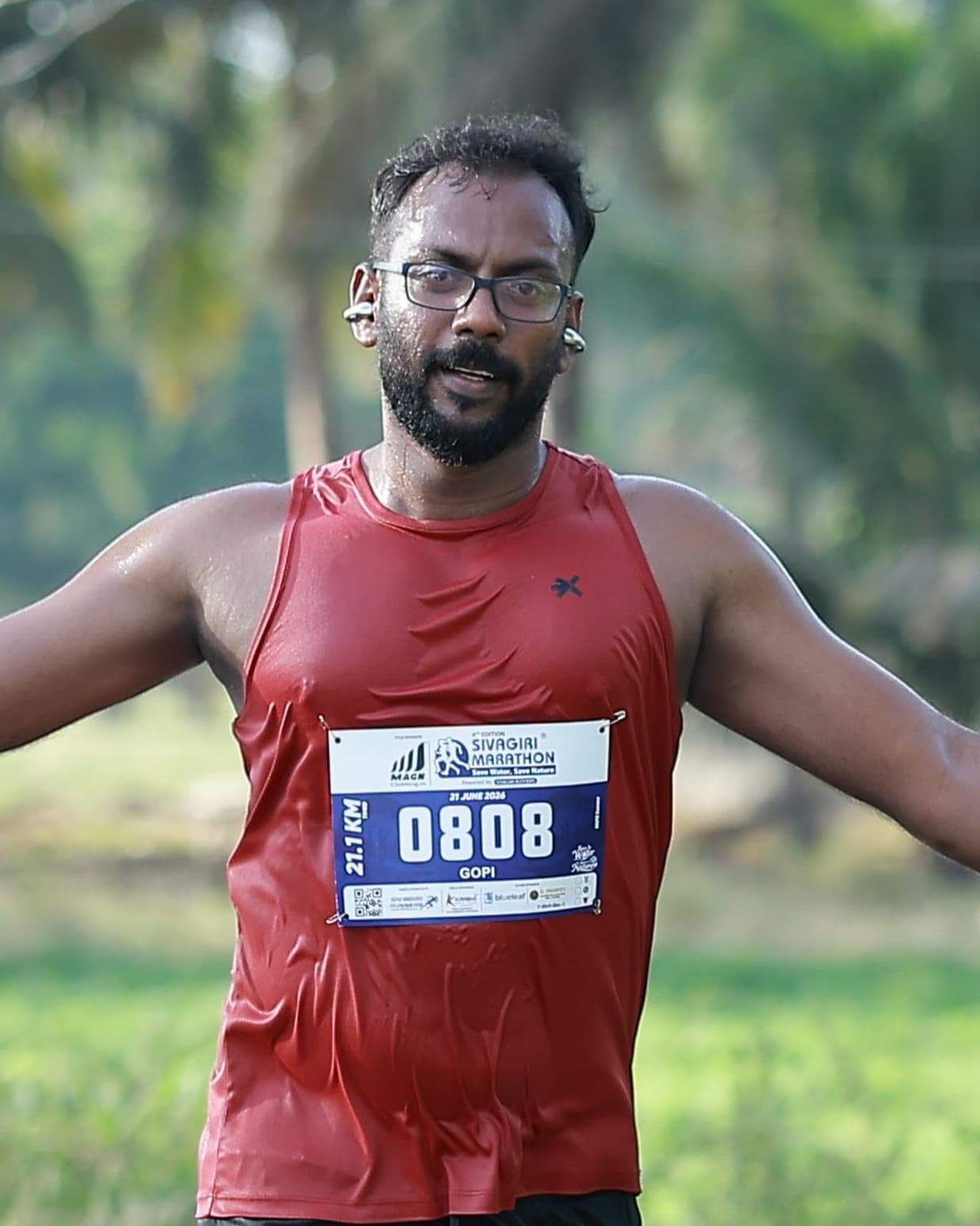 Gobikrishnanraja running a half marathon on a palm-lined road in a red vest, arms spread wide.