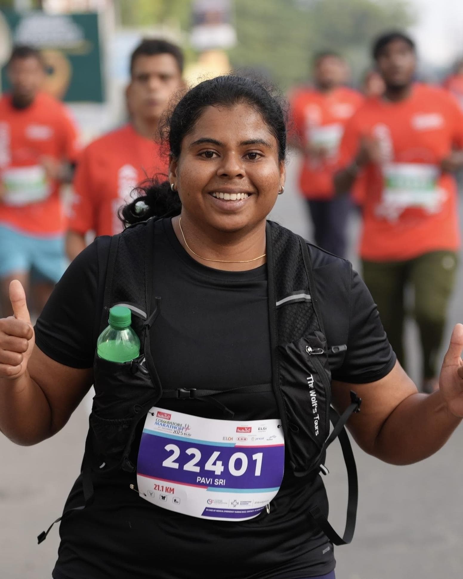 Pavithra Sri mid-race in a black running vest and hydration pack, both thumbs up and smiling at the camera, with other runners behind her.