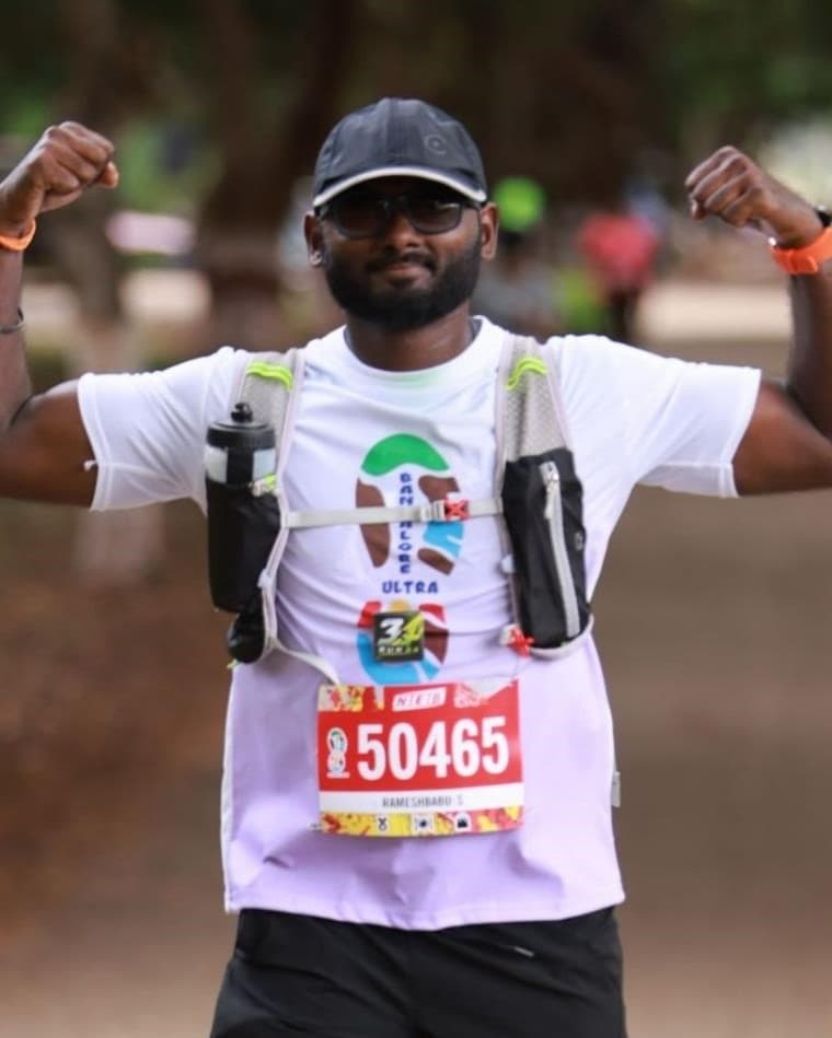Ramesh Babu running an ultra in a RUN 34 tee and hydration vest, cap and sunglasses on, flexing both arms at the camera.